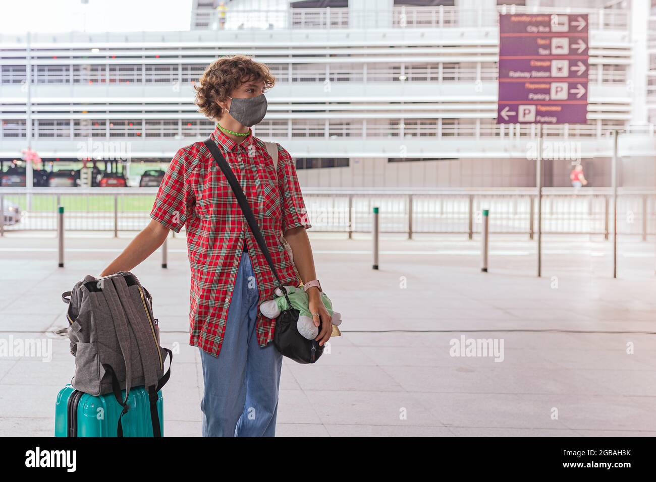 Portrait of attractive teenage girl with curly hair in protective mask with luggage for travel on blurred background of airport building Stock Photo