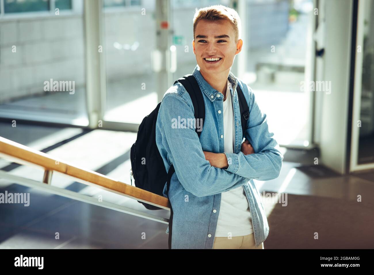 Boy in casual wear and arms crossed leaning to stairs in college. Student standing in college looking away and smiling. Stock Photo