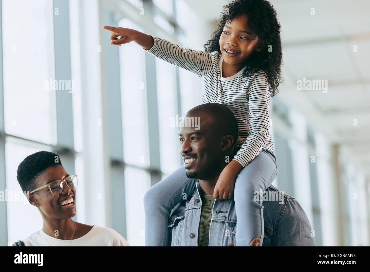 Girl on shoulder of father showing airplanes while walking with mother at airport. Father carrying young daughter on shoulder and walking with wife at Stock Photo