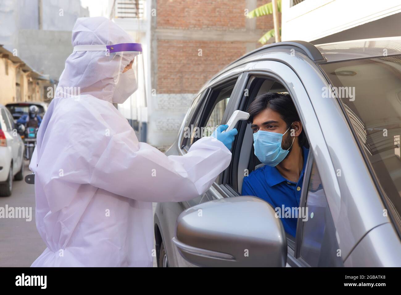 A health worker in PPE kit taking temperature of a man in car with thermometer gun. Stock Photo