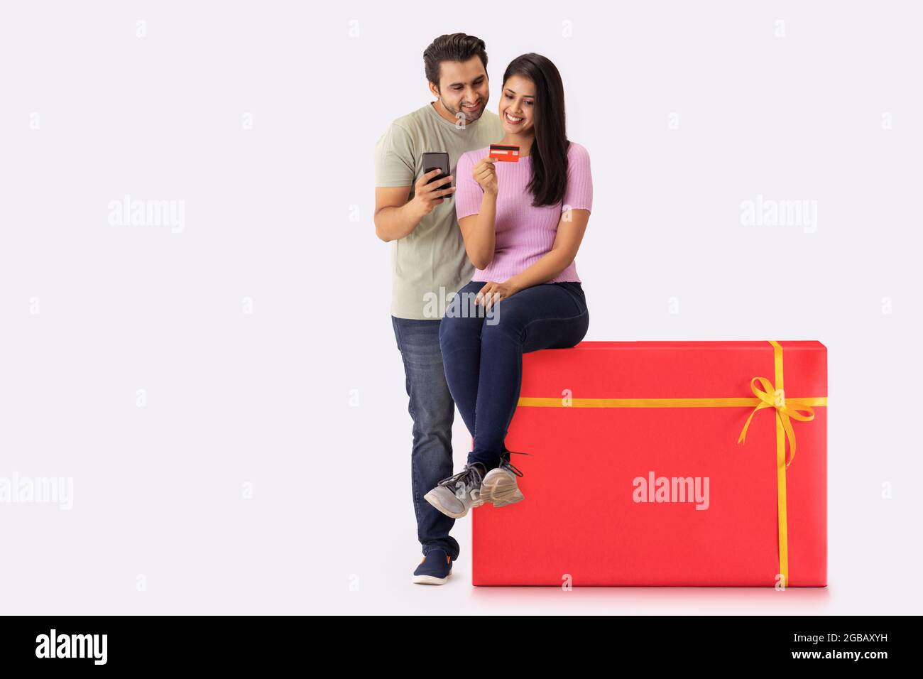 A young woman sitting on a gift shaped stool with a man beside,making online payment with credit card and phone. Stock Photo