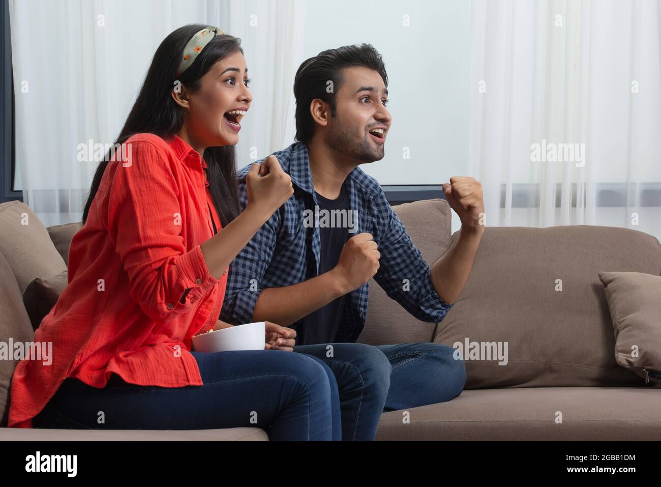 A young couple cheering for a team excitedly sitting in a room. Stock Photo