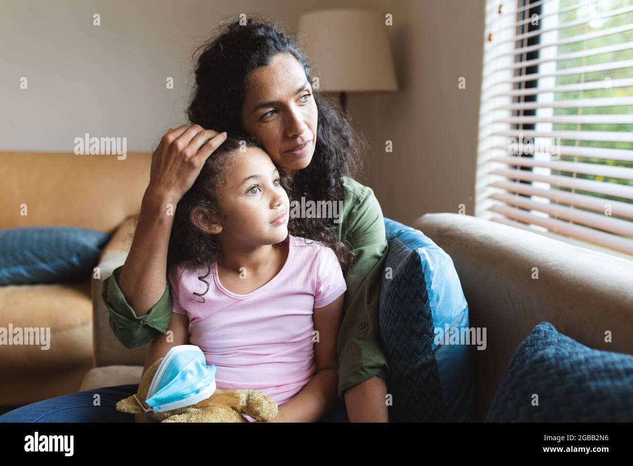 Mixed race mother and daughter sitting on sofa, embracing and looking through window Stock Photo