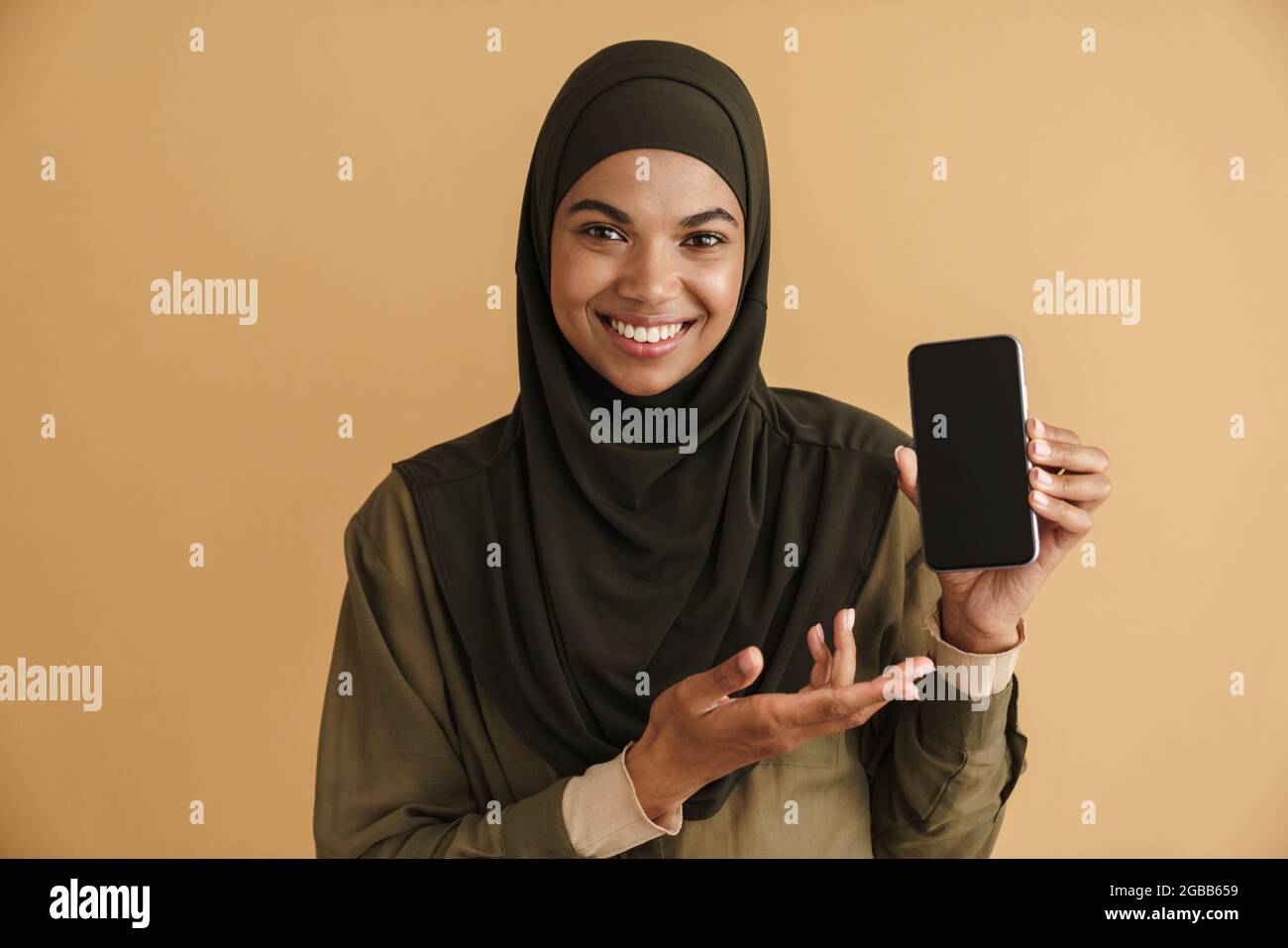 Black muslim woman in hijab smiling and showing mobile phone isolated over beige background Stock Photo