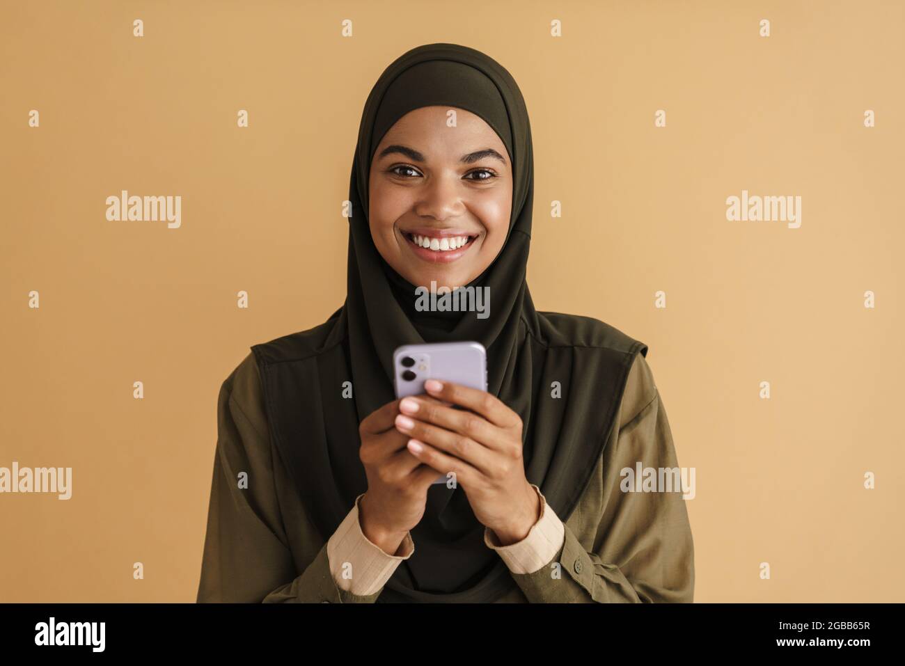 Black muslim woman in hijab smiling and using mobile phone isolated over beige background Stock Photo