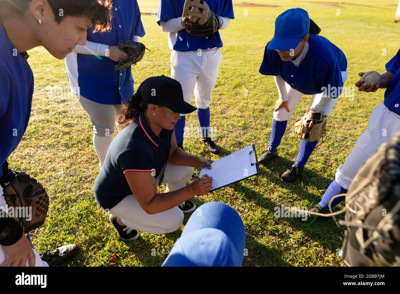 Diverse group of female baseball players in huddle around squatting female coach with clipboard Stock Photo
