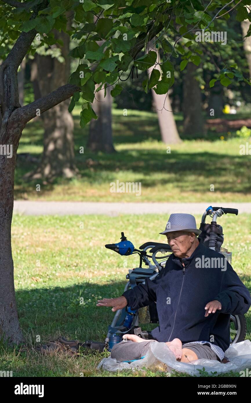 A seated 86 year old man does Falun Gong slow-moving exercises close to a tree. In a park in Queens, New York, a very diverse place. Stock Photo