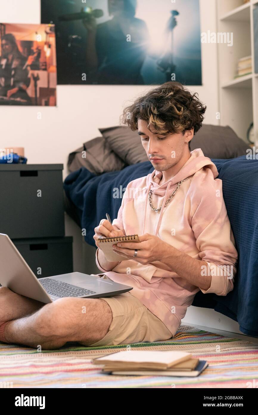 Teenage guy making notes in notepad while sitting on the floor by his bed Stock Photo