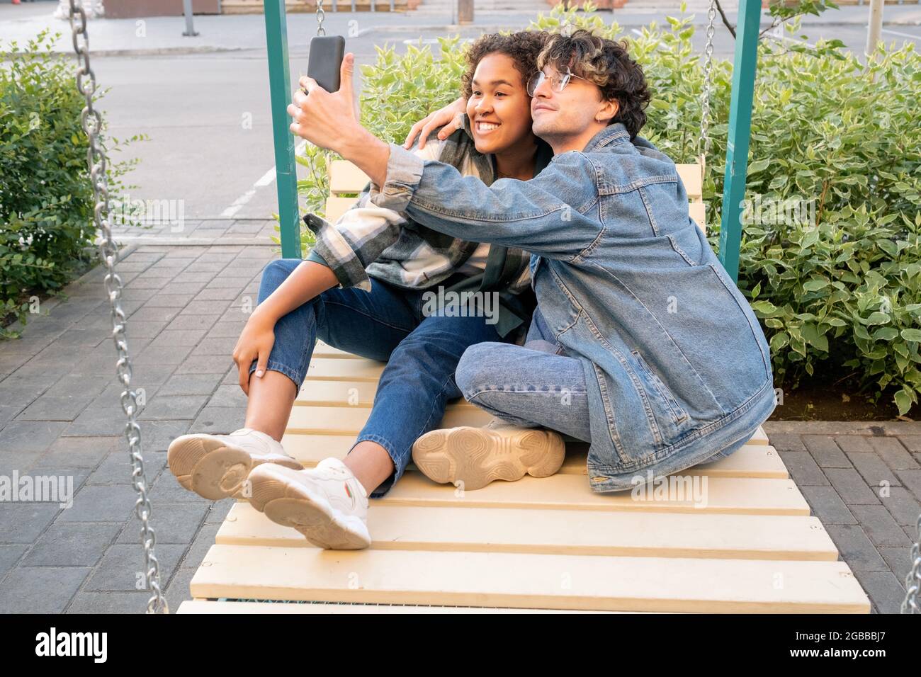Carefree teenage dates making selfie on swings while guy embracing his happy girlfriend Stock Photo
