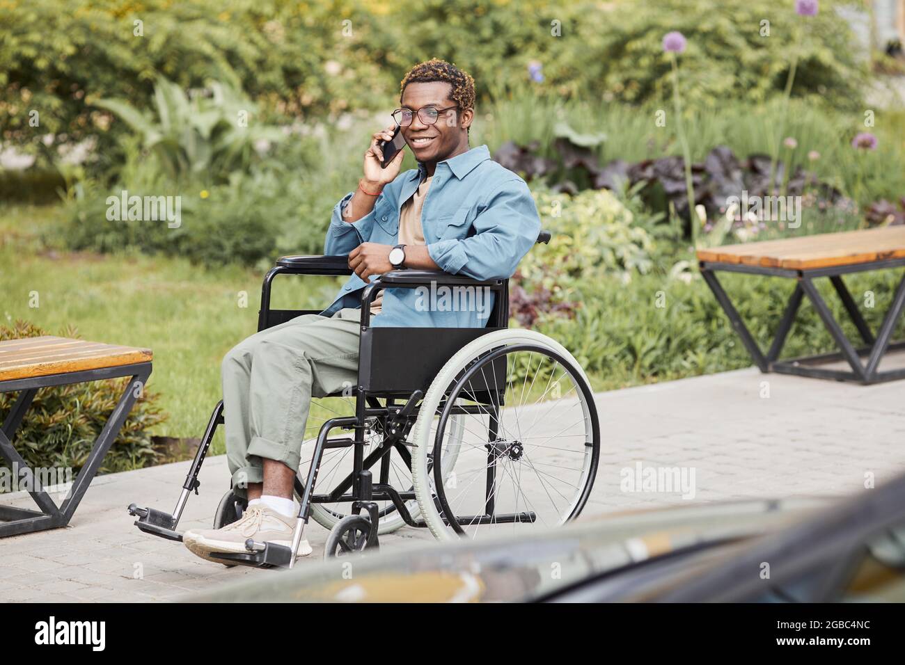 Happy handsome disabled young African-American man in eyeglasses sitting in wheelchair and talking by phone Stock Photo