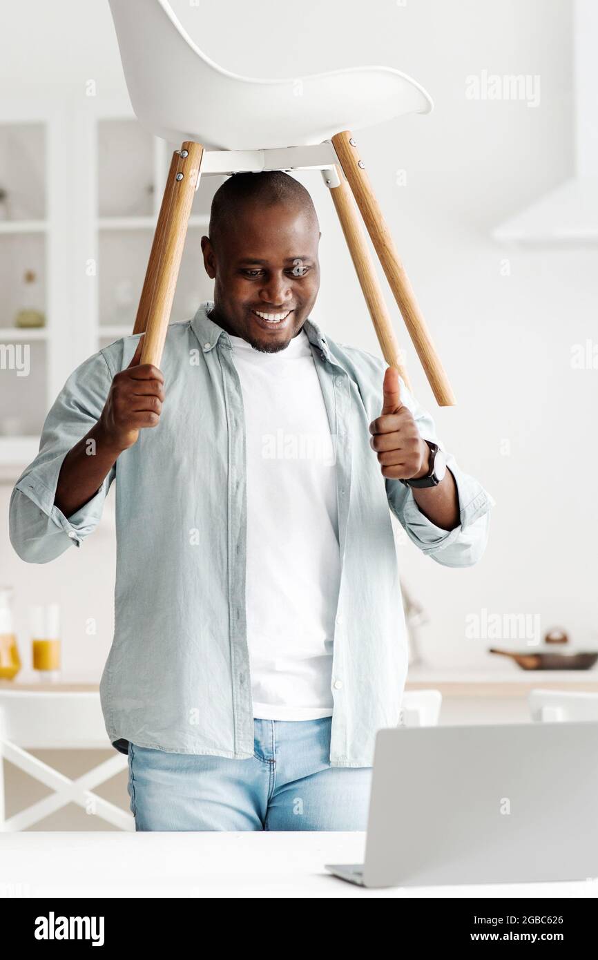 Funny video about renovation and furniture. Happy black man showing  ready-made chair, having video chat on laptop Stock Photo - Alamy