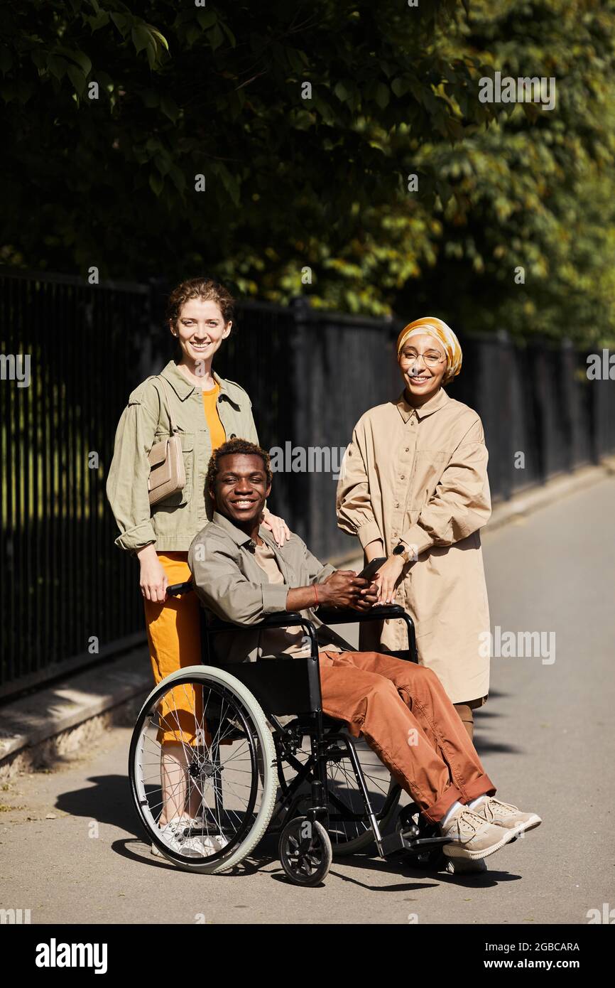 Portrait of smiling young women standing near handsome handicapped man in wheelchair outdoors Stock Photo