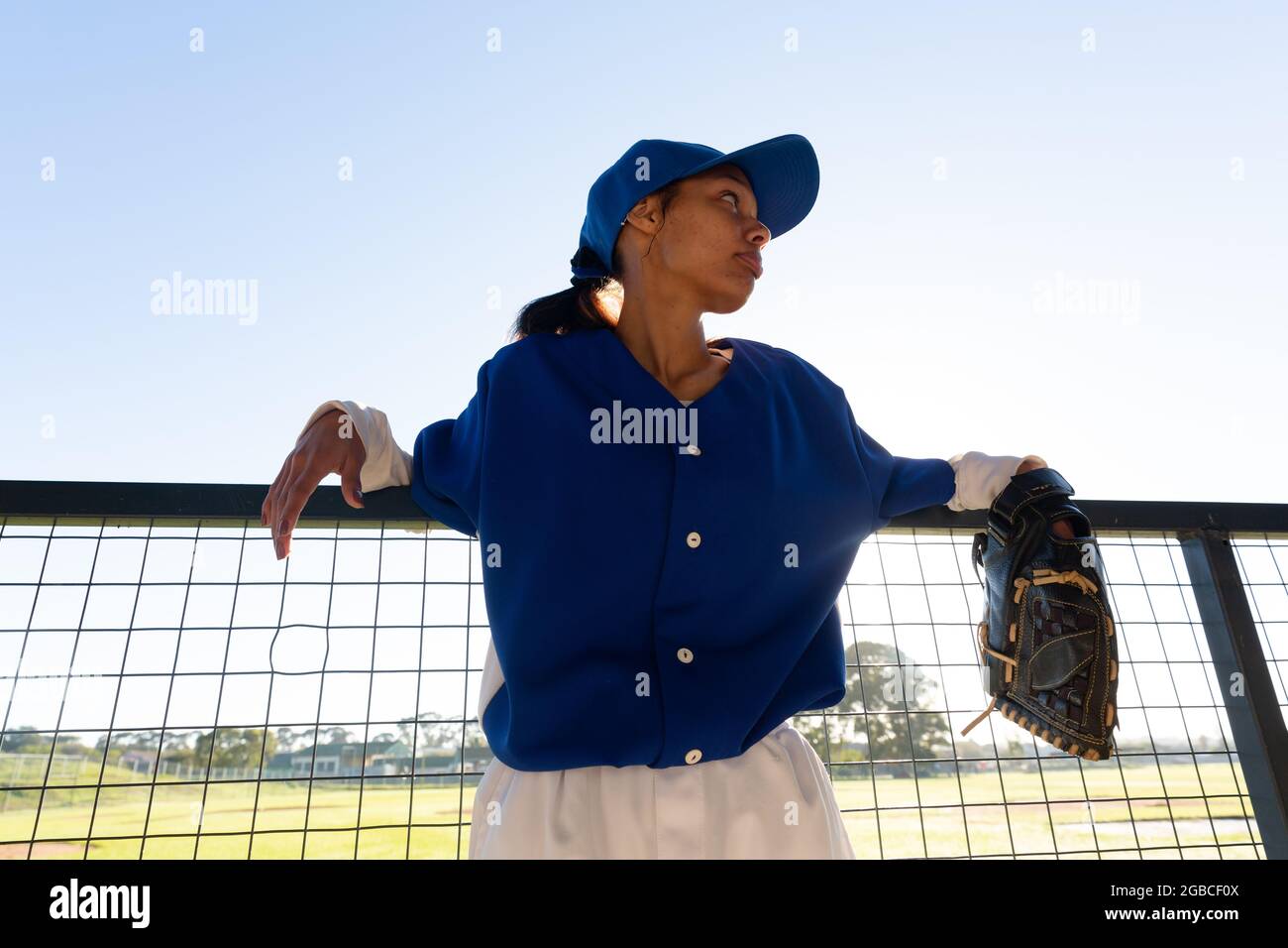 Mixed race female baseball player wearing glove, leaning on fence in