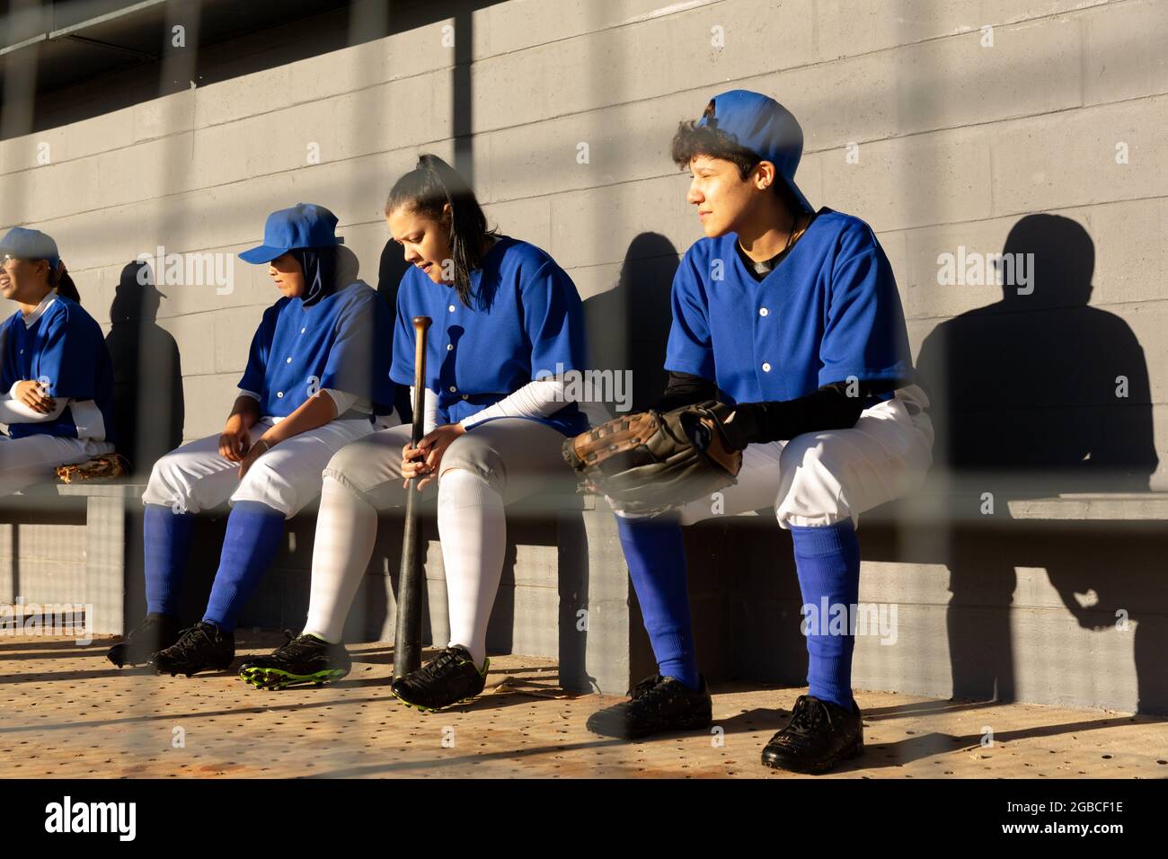 Diverse group of female baseball players sitting on bench in sun, waiting to play Stock Photo