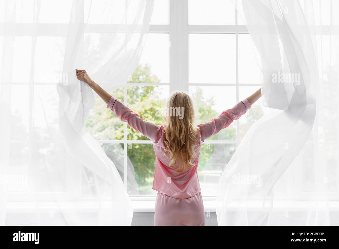 Young pretty blonde woman in pink pajamas woke up, look at large panoramic window in bedroom Stock Photo