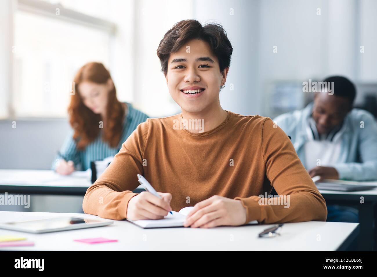 Asian male student sitting at desk writing in classroom Stock Photo