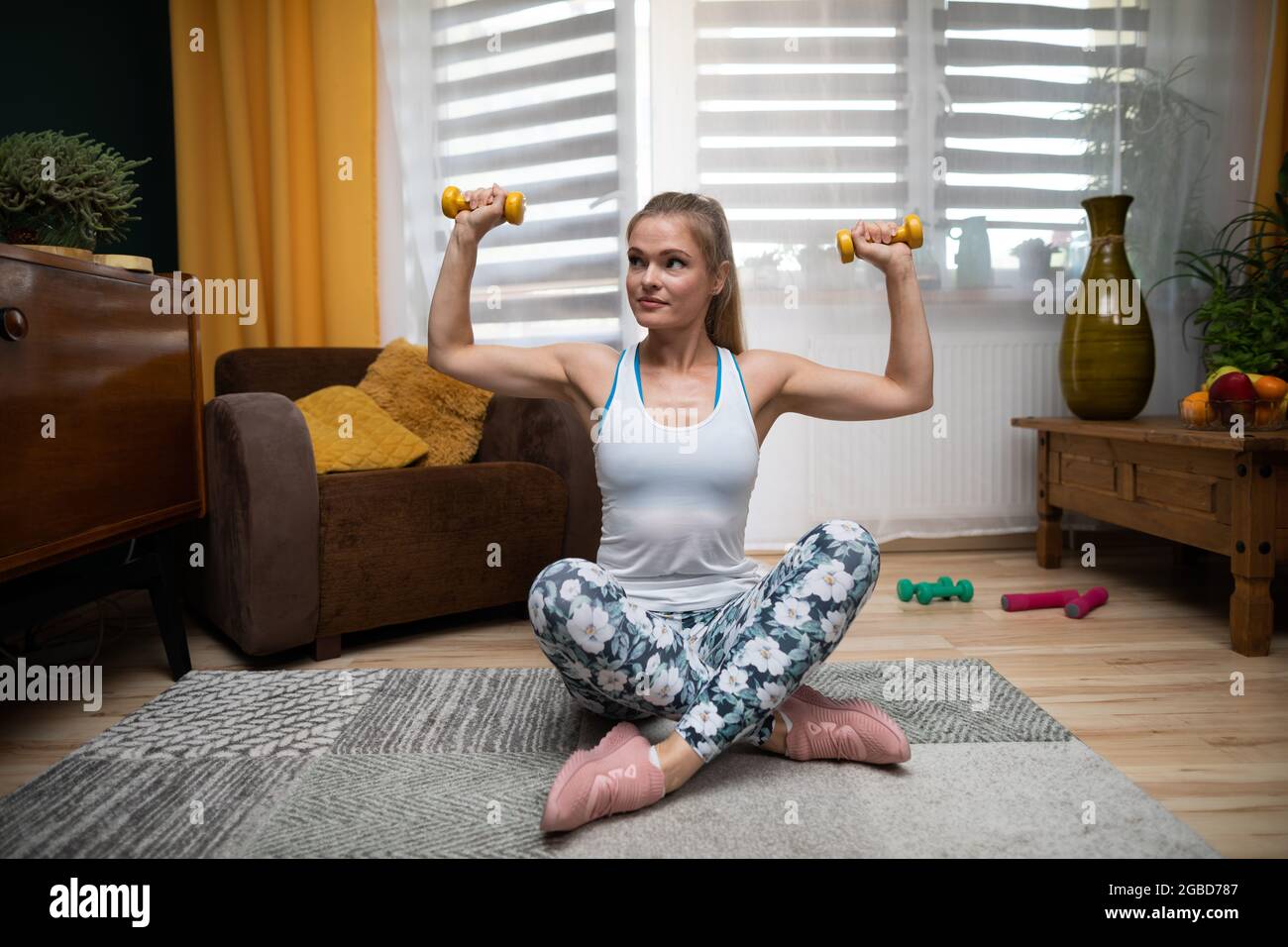 A woman sits cross-legged and holds weights in both hands and exercises with them. Fitness at home. Stock Photo