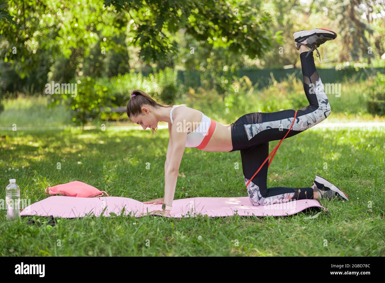 Sportswoman does back leg lift with elastic band kneeling on lawn grass Stock Photo