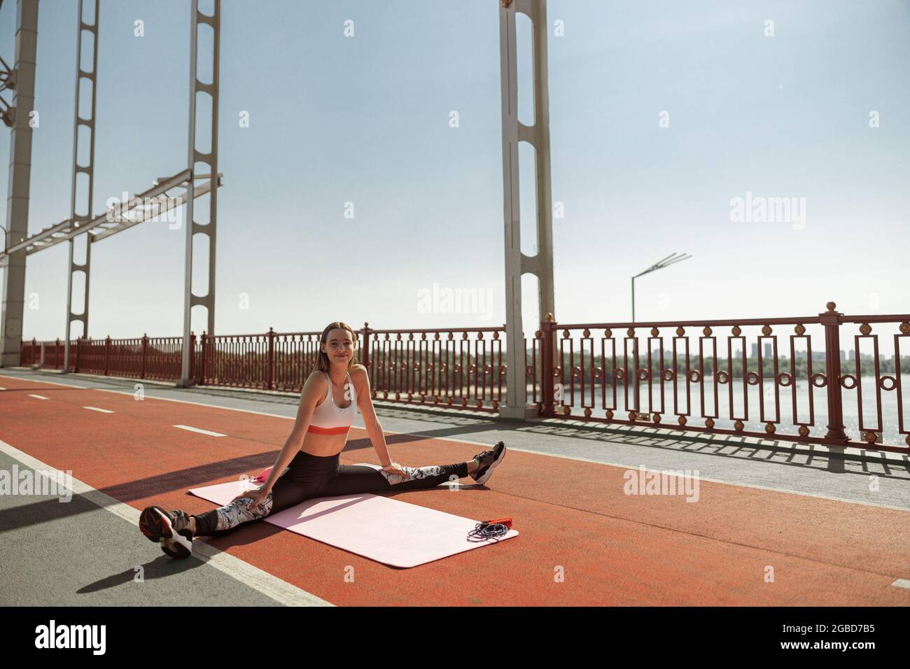 Attractive positive woman does stretching exercise sitting on contemporary bridge Stock Photo