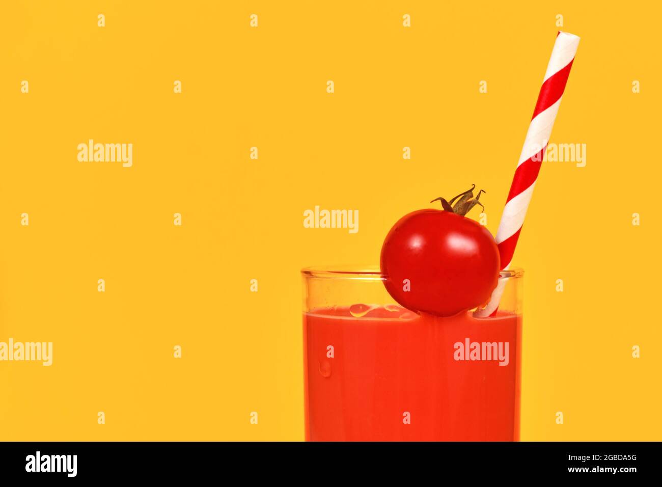 Glass with red tomato juice, cherry tomato and striped drinking straw on side of yellow background with copy space Stock Photo