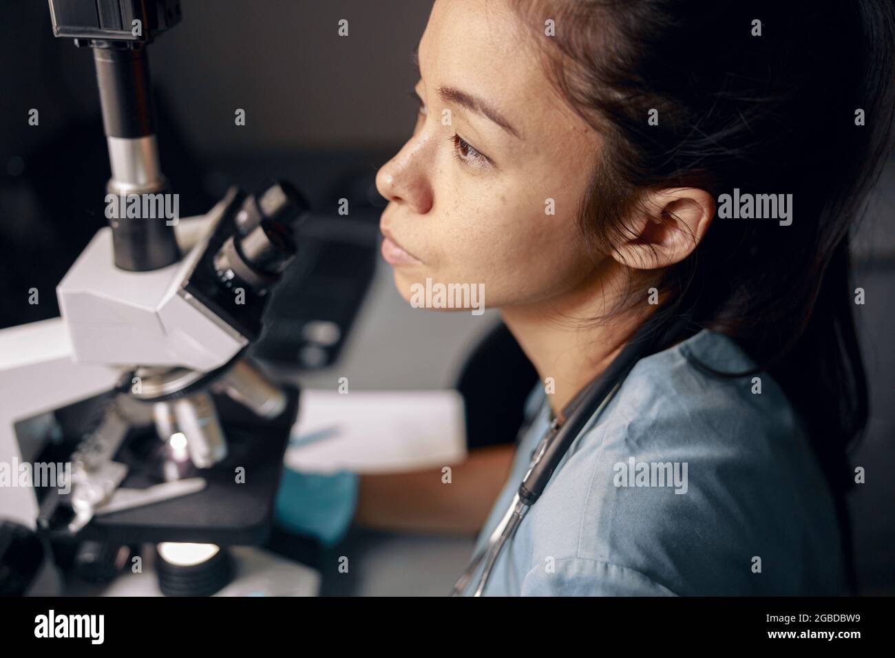 Thoughtful Asian woman in uniform with modern microscope at workplace in hospital laboratory Stock Photo
