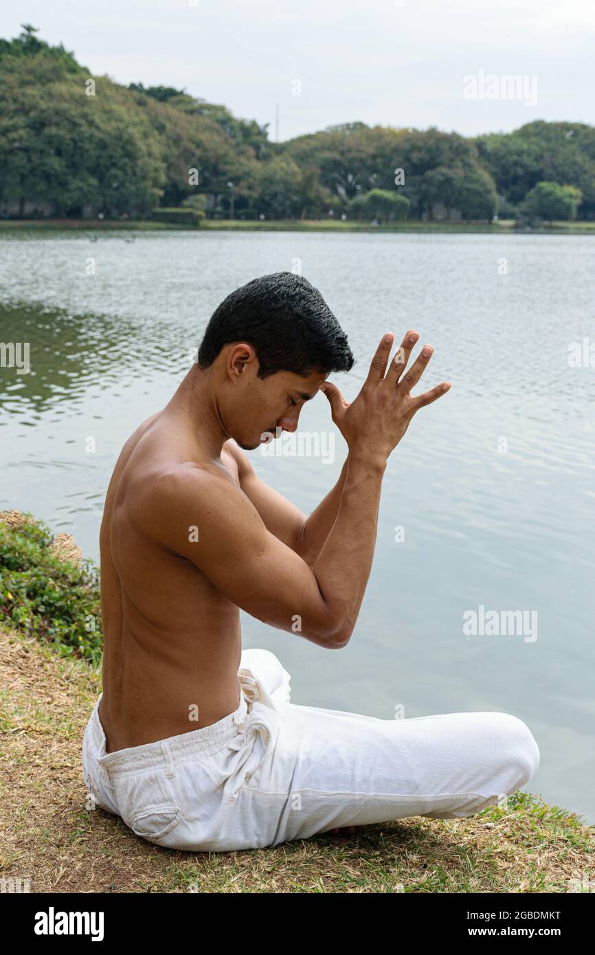Brazilian young man sitting on the edge of a lake, with his hands opposite and thumbs touching his forehead (side view).. Stock Photo