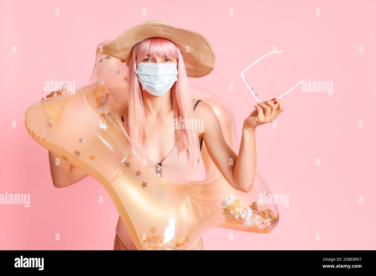Woman in straw hat and swimsuit with inflatable rubber ring wearing hygienic face mask to prevent contagious coronavirus on resort beach, posing Stock Photo