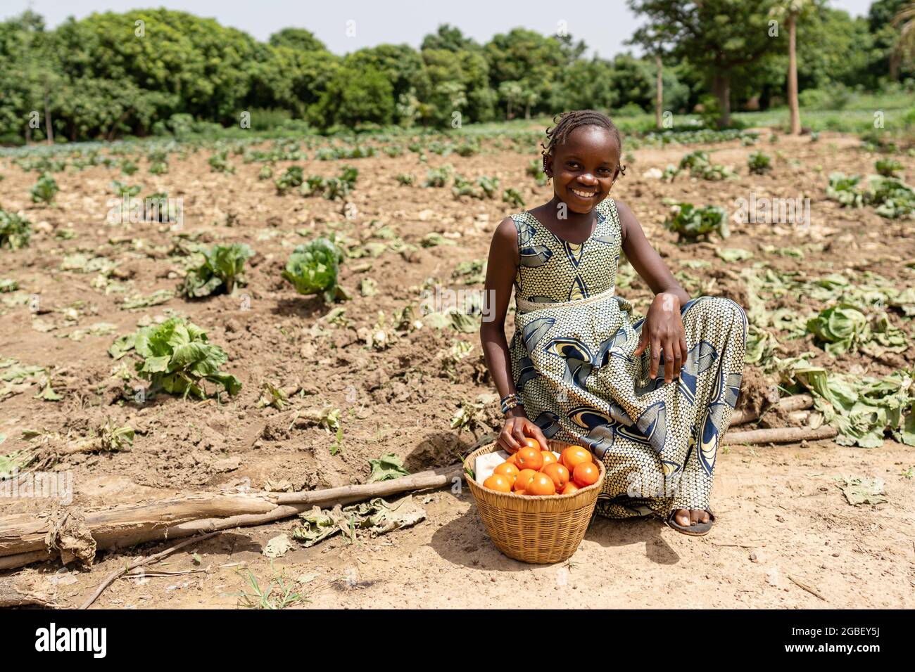 In this image, a smiling African girl is resting on the long way to the market under the hot sun, sitting on the border of a cabbage field with her fu Stock Photo