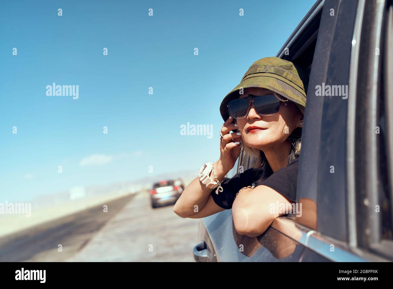 asian woman traveler with hat and sunglasses sticking head out of rear window of car looking at view Stock Photo