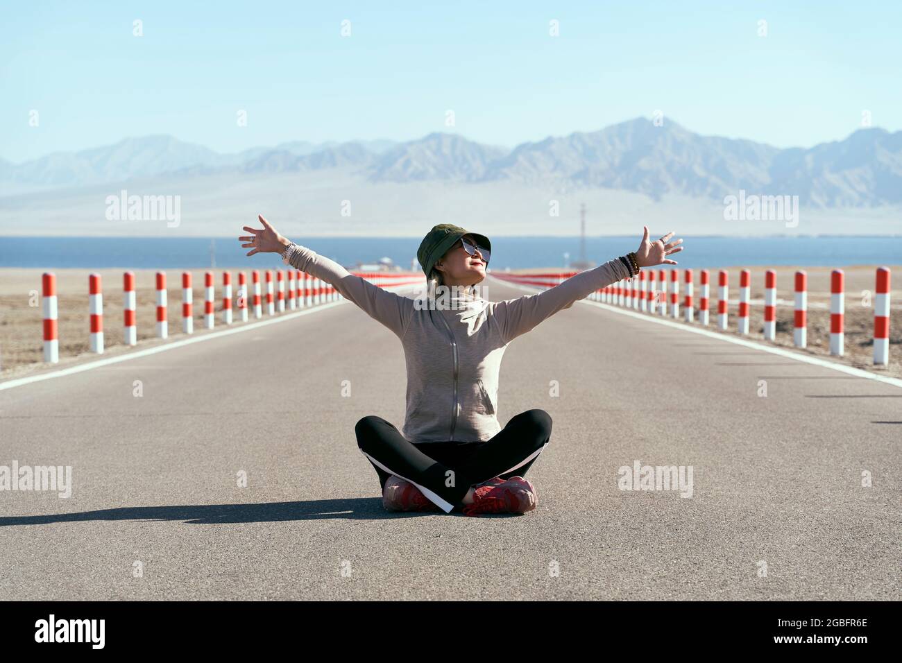 asian woman tourist sitting in the middle of an empty open road with lake and rolling mountains in background, leg crossed arms outstretched. Stock Photo