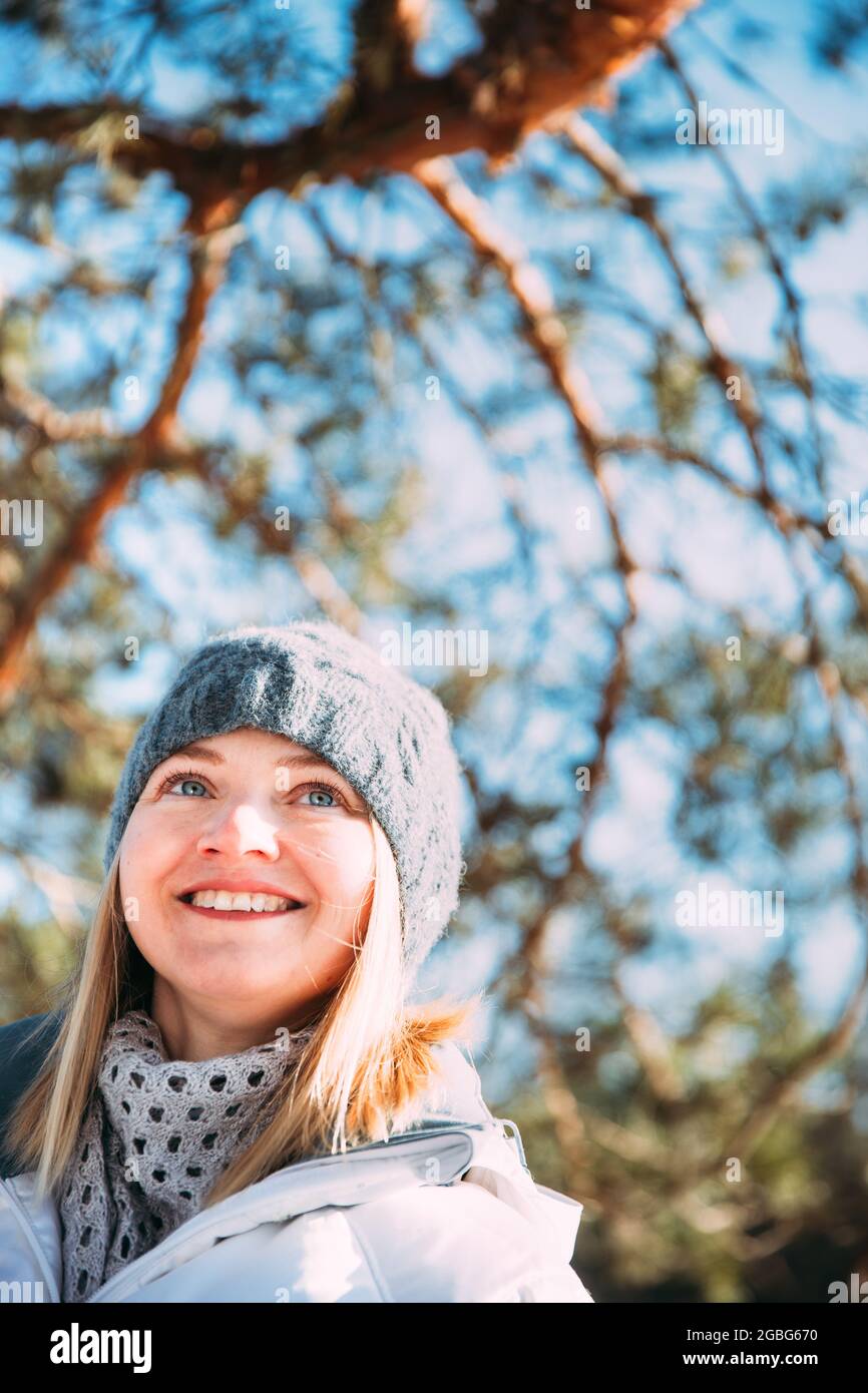 Young Caucasian Lady Woman Dressed In White Jacket And Grey-blue, Gray Hat Posing Near Pine Tree. Close Up Portrait. beautiful young woman smiling Stock Photo
