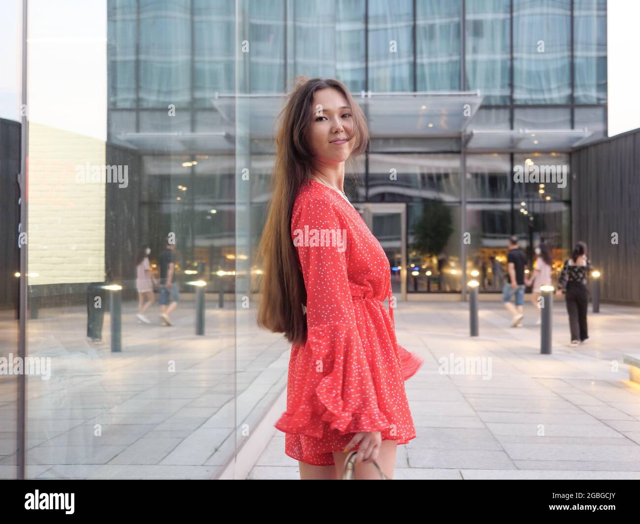 Attractive smiling Asian girl in red dress smiles next to glass facades ...