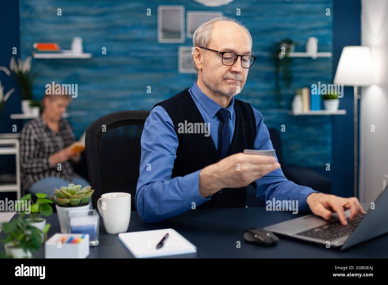 Senior man using credit card to check bank account. Elderly man checking online banking to make shppping payment looking at laptop while wife is reading a book sitting on sofa. Stock Photo