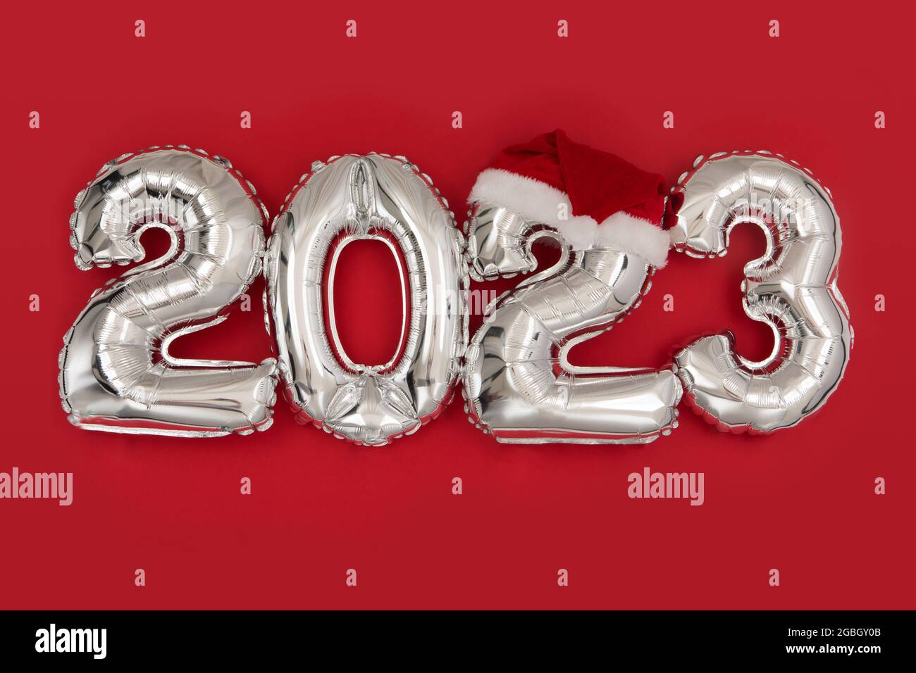 2023 inflatable balloons on red background with santa hat. Happy New year 2023 celebration. Silver foil balloons numeral 2023. Postcards and posters C Stock Photo