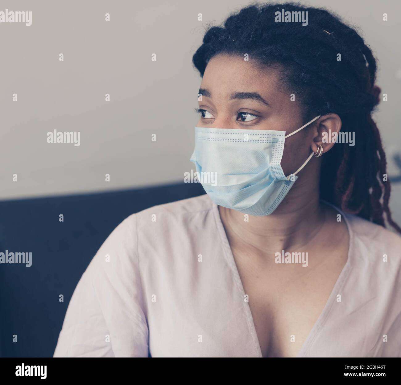 Young African American woman with a medical mask on her face sits in a chair on a light background and looks anxiously to the side. Color graded Stock Photo
