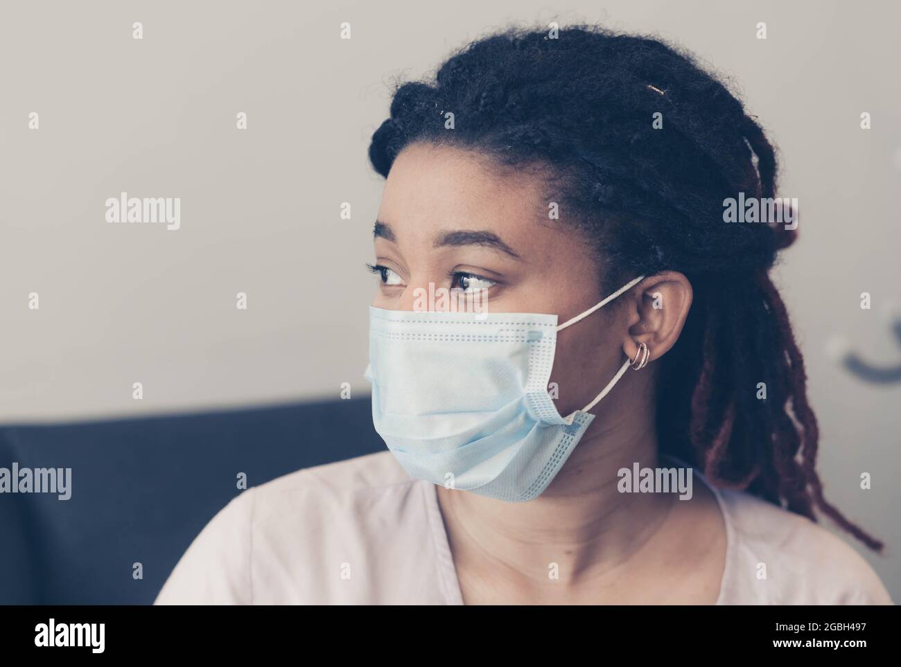 A woman of African American appearance sits down against the background of a light wall. On the face of a person a protective medical mask. No emotion Stock Photo