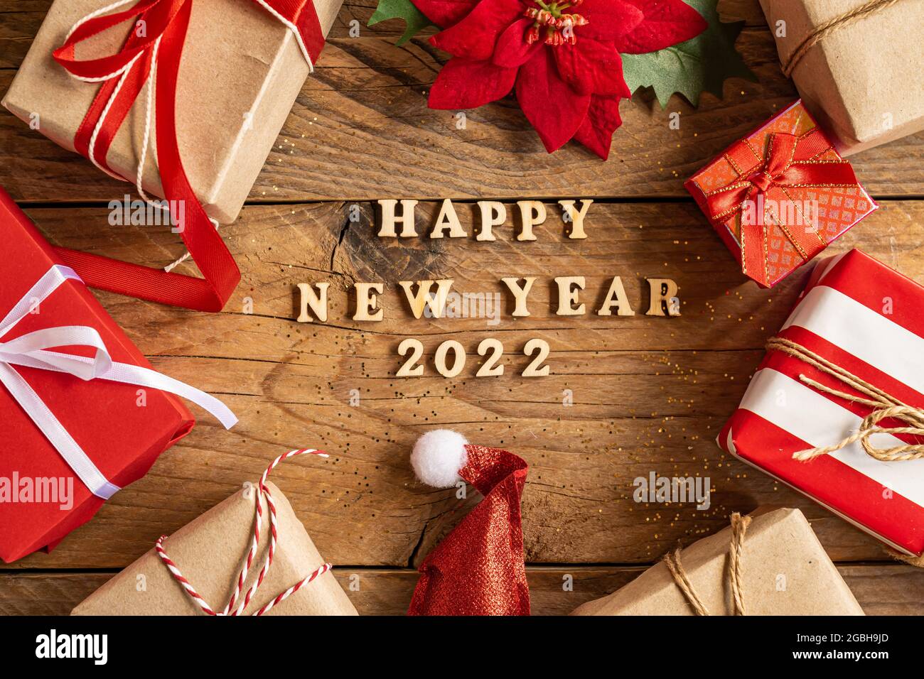 happy new year 2022 festive background with wooden letters and craft gift boxes, poinsettia and santa hat on wooden background. Stock Photo