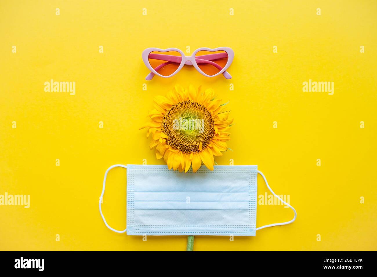 Funny face made of pink heart-shaped sunglasses, a protective medical mask and a sunflower on a bright yellow background Stock Photo