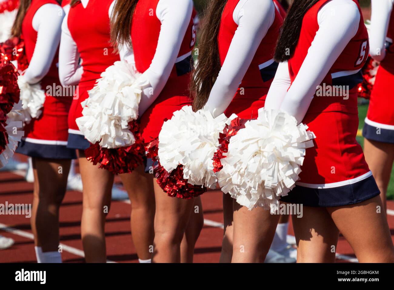 Rear view of high school cheelearders standing in a line together holding their pop poms behind them while watching the football game. Stock Photo
