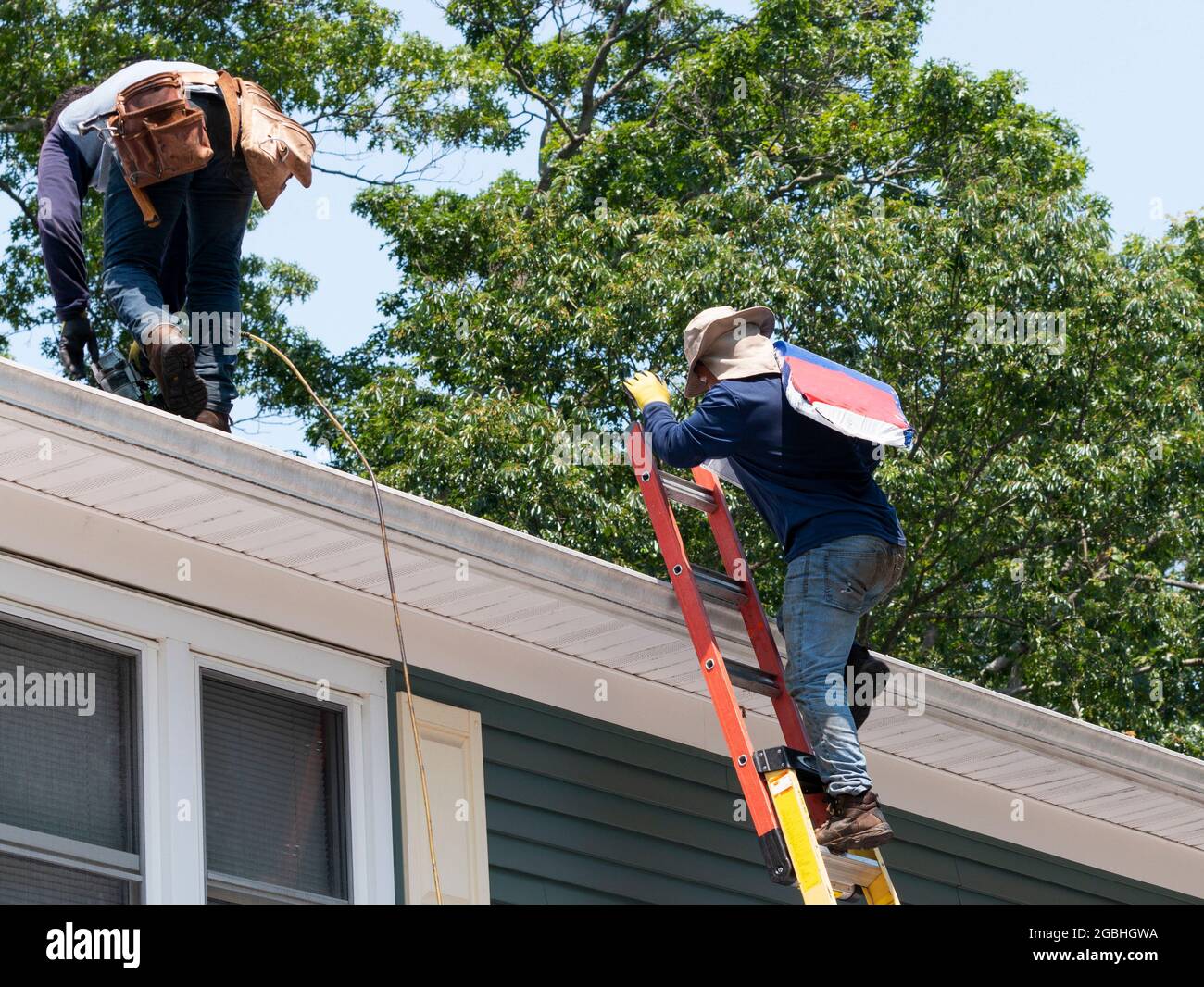 contractor climbing ladder with roof shingles while another nail the shingles on a residential house. Stock Photo