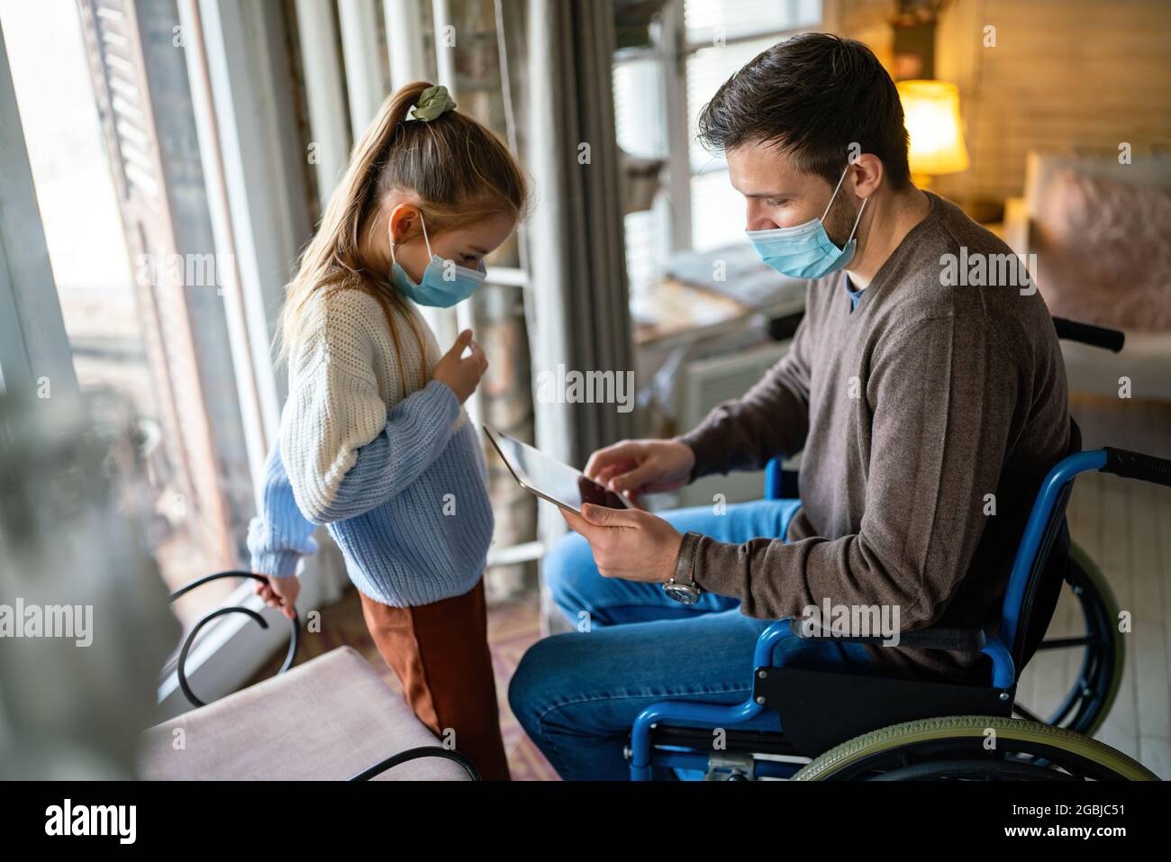Father with disability in wheelchair using tablet at home with child while wearing masks. Stock Photo