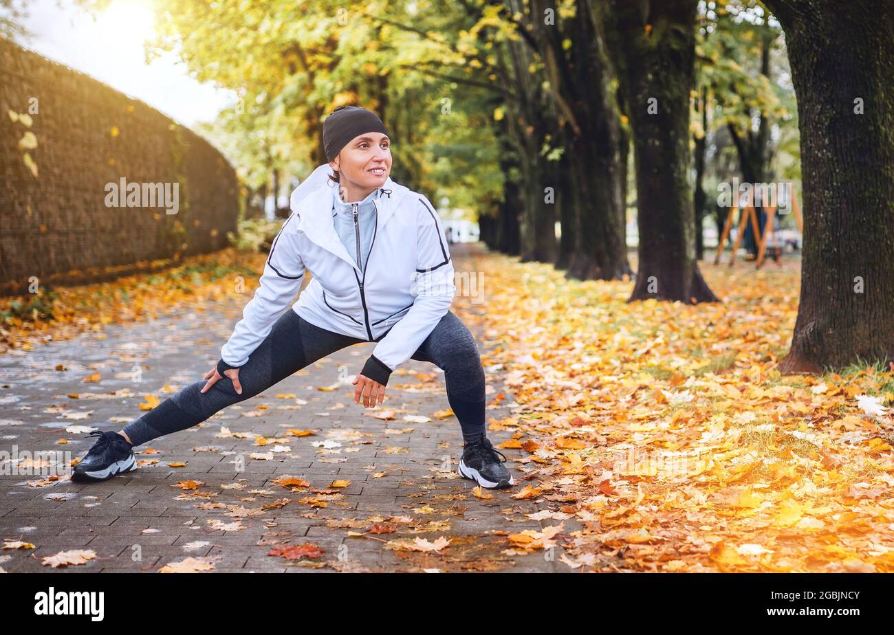 Fit athletic woman doing workout before jogging in autumnal city park on the kids playground. Young fitness female runner stretching legs while warmin Stock Photo
