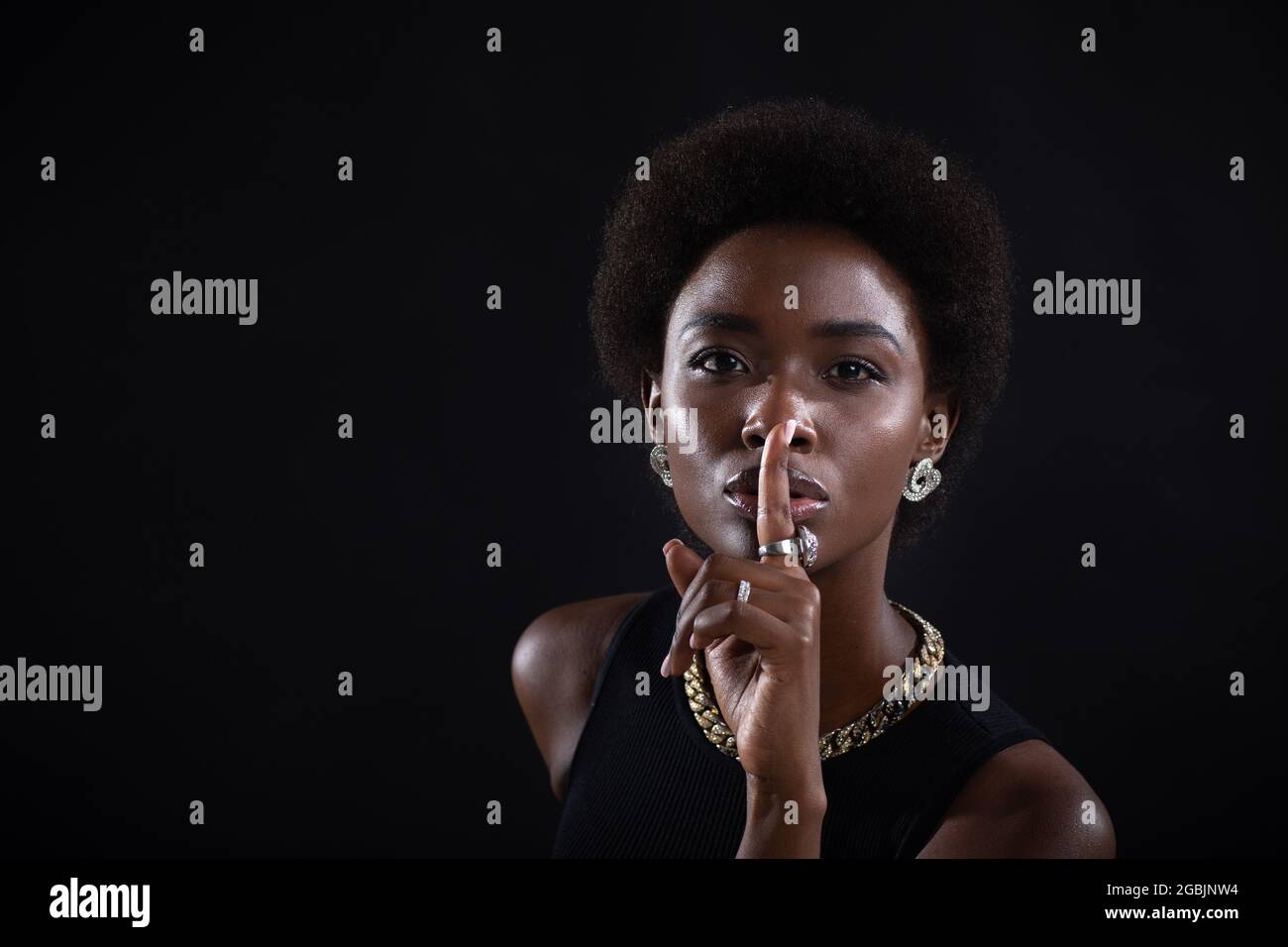 Closeup of beautiful young african american dark-skinned woman with finger on her lips showing sh silence gesture on black background. Stock Photo
