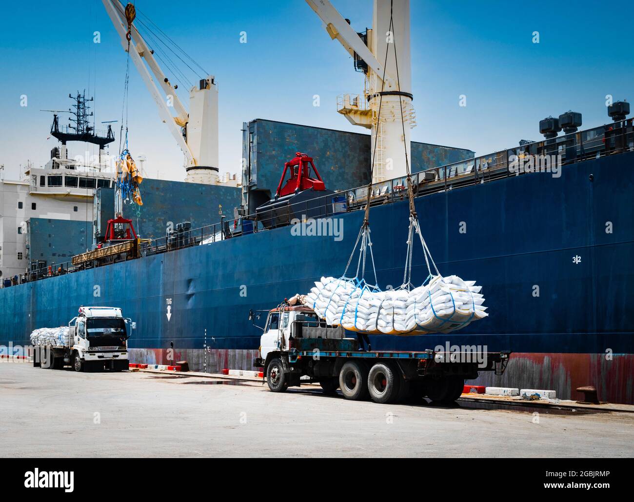 Ship crane lift-off slings of sugar bags cargo from truck and load into ship hold at seaport terminal for export. Stock Photo