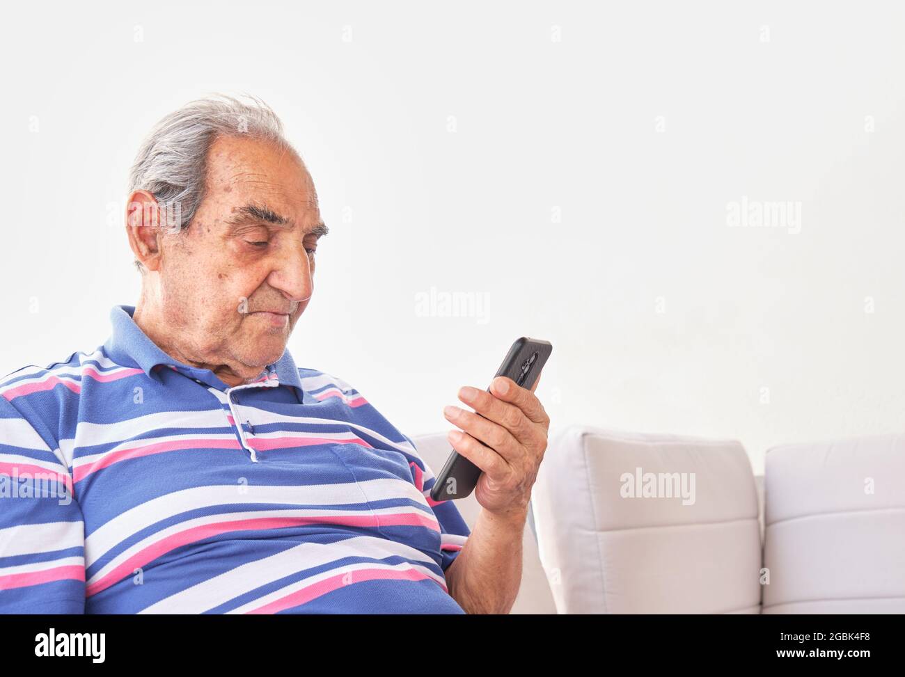 older dark-haired man with gray hair in striped shirt sitting holding a smartphone with light background Stock Photo