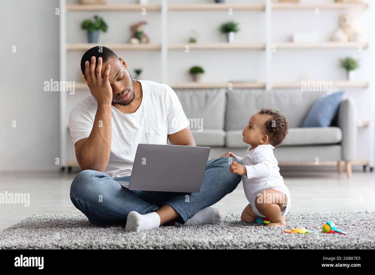 Stressed Black Man Trying To Work With Laptop While Baby Distracting Him Stock Photo