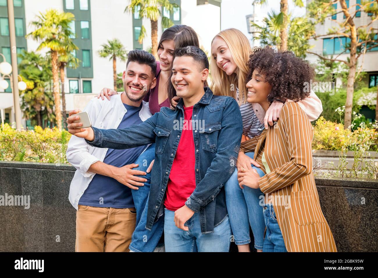 Group of five happy multicultural friends taking a selfie on a mobile phone on a city street laughing and smiling as they pose for the camera Stock Photo