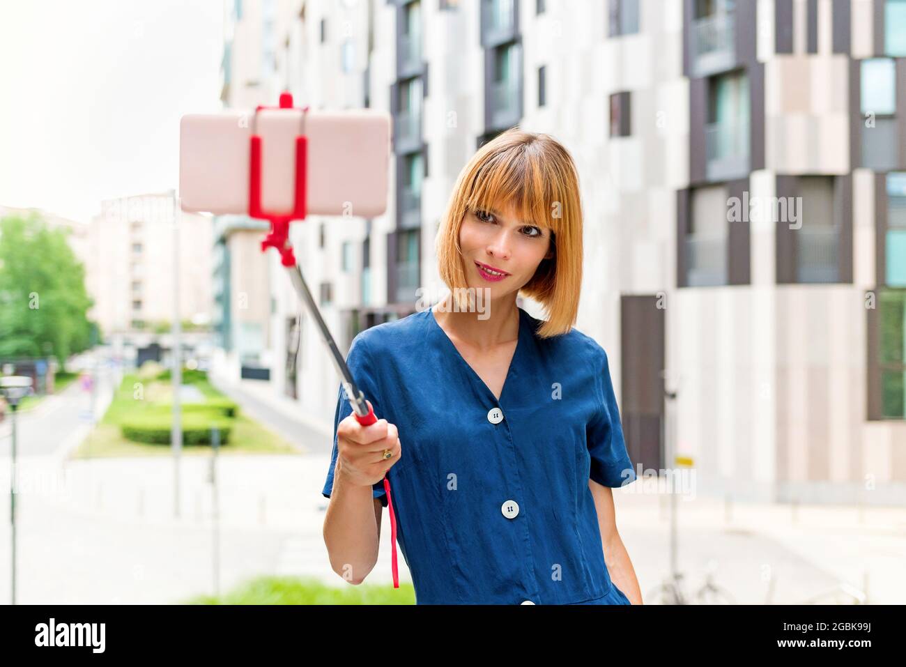 Slender attractive woman taking a selfie on a city street using her mobile phone on a selfie stick as she smiles quietly for the camera in a high key Stock Photo