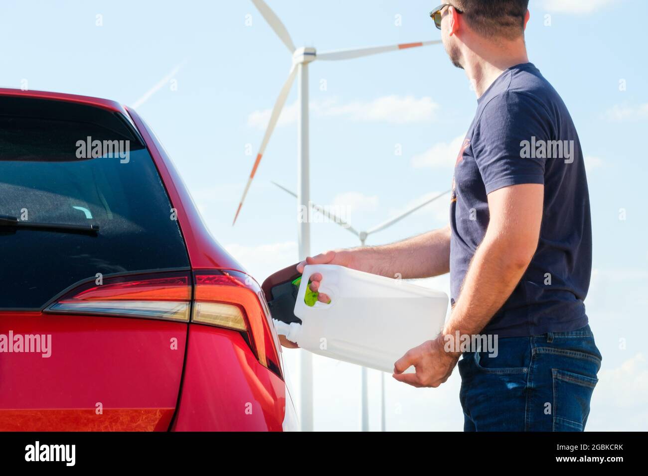 Man filling a diesel engine fluid from canister into the tank of red car on the background of wind turbines. Reduction of air pollution and Stock Photo