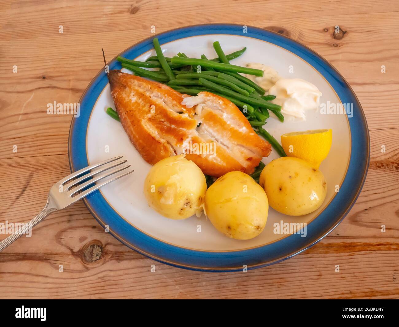 A  lunch of Smoked Haddock  with green beans new potatoes a slice of lemon mayonaise and tartare sauce on blue edged white plate on a wooden table Stock Photo